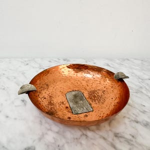 May include: A hammered copper bowl with a tarnished finish. The bowl has two silver-colored handles and a rectangular metal label. The bowl is round and has a shallow depth. The copper has a reddish-brown color.