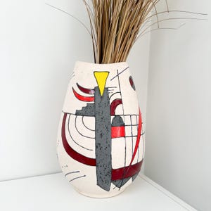 May include: Cream-colored ceramic vase with abstract geometric design in red, gray, yellow, and black. The vase has a tall, oval shape and contains dried ornamental grass. The design features lines, shapes, and patterns.