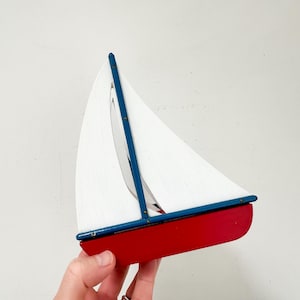 May include: A wooden sailboat with a red hull, white sails, and blue trim. The boat is held by a hand and features brass-colored rivets. The design is reminiscent of nautical decor.