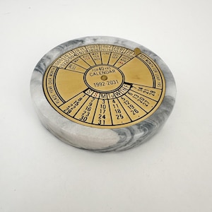 May include: A round, marble calendar with a brass dial. The dial displays months, days, and years from 1992 to 2031. The marble base is gray and white, and the brass dial has black lettering. The calendar is designed to last for 40 years.