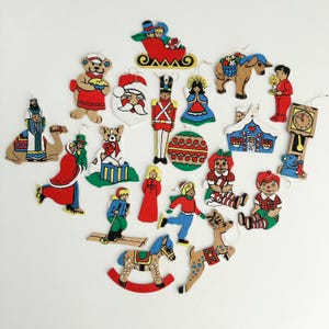 May include: A selection of vintage Christmas tree decorations in diverse shapes. The ornaments feature figures such as Santa Claus, angels, toy soldiers, and children, all with a rustic, hand-painted look. The colour scheme is dominated by red, green, and brown.