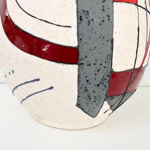 May include: A ceramic vase with a cream-colored base, featuring geometric designs in red, gray, and black. The vase has a rounded shape with a textured surface and abstract patterns.