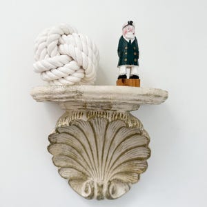 May include: A white wall-mounted shelf shaped like a seashell with a distressed finish. On the shelf sits a white monkey fist rope knot and a small hand-painted wooden figurine of a sea captain wearing a dark green coat and white hat.