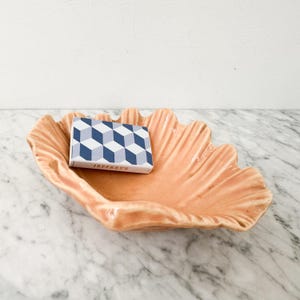 May include: A peach-colored ceramic bowl shaped like a large clam shell with a glossy finish. Resting inside the bowl is a rectangular matchbox featuring a blue, white, and grey geometric cube pattern and the text JEFFREY'S printed on the side.