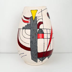May include: Cream-colored ceramic vase with an abstract design. The vase features a gray, textured vertical element, red curved lines, a yellow triangle, and black geometric shapes. The vase has a rounded, elongated shape.