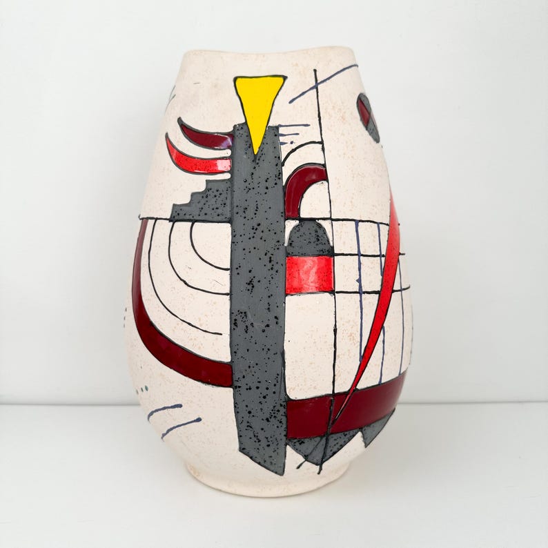 May include: Cream-colored ceramic vase with an abstract design. The vase features a gray, textured vertical element, red curved lines, a yellow triangle, and black geometric shapes. The vase has a rounded, elongated shape.