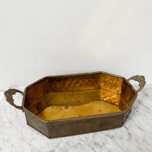 May include: A vintage brass planter with an octagonal shape and decorative handles. The interior has a golden hue, and the exterior shows signs of age. The planter is approximately 12 inches long and 6 inches wide.