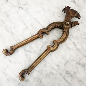 May include: A vintage-style brass nutcracker featuring a detailed bird-shaped head at the top. The tool has two long, curved handles with decorative etched patterns and a serrated inner grip area for cracking nuts, finished in a dark, aged patina.