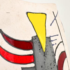 May include: Close-up of a ceramic vase with an abstract design. The vase features a cream-colored background with bold geometric shapes in red, yellow, and gray, outlined in black. The design includes a yellow triangle and red curved lines.