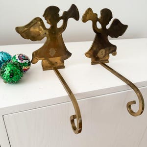 May include: A pair of brass stocking holders featuring silhouettes of angels playing trumpets. Each holder has a flat vertical angel figure attached to a long horizontal arm that ends in a curved hook. Three sequined ornaments are visible nearby.