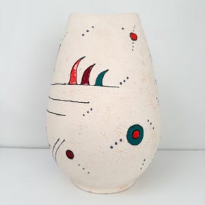 May include: Cream-colored ceramic vase with abstract red, green, and black painted details. The vase has a textured surface and a rounded shape, with a series of dots and lines decorating the exterior.