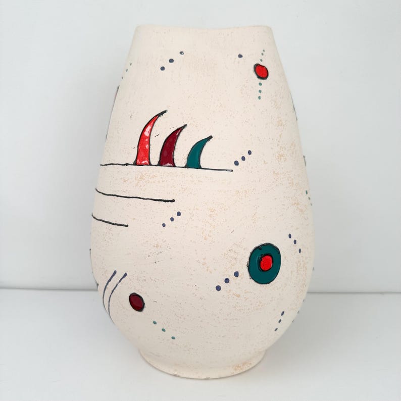 May include: Cream-colored ceramic vase with abstract red, green, and black painted details. The vase has a textured surface and a rounded shape, with a series of dots and lines decorating the exterior.