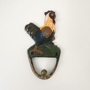 May include: A cast iron wall hook featuring a painted rooster standing on a green grassy base. The rooster has yellow, brown, and blue plumage with a red comb. The hook below is finished in a distressed bronze tone with two mounting holes.