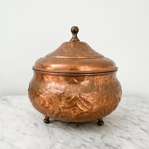 May include: A round copper container with a matching lid and a small brass knob handle. The exterior features an embossed pattern across the body and lid. The vessel is supported by three small, rounded feet, showcasing a metallic finish.