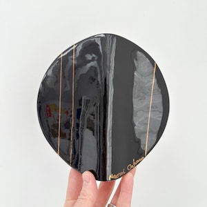 May include: A black, oval decorative plate with copper-coloured vertical stripes. The plate has a glossy finish and is held by a hand. The artist's signature, "Naomi Cahane," is visible on the bottom right.