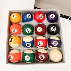 May include: A set of sixteen billiard balls arranged in a square cardboard box. The collection includes solid-colored balls numbered one through eight and striped balls numbered nine through fifteen, plus one solid white cue ball with no number.