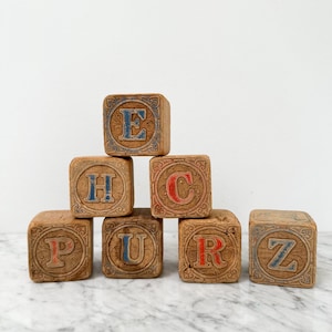 May include: Seven square wooden blocks arranged in a pyramid shape. Each block features an embossed letter surrounded by intricate scrollwork patterns. The letters E, H, U, and Z are painted in blue, while the letters C, P, and R are in red.