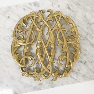 May include: A gold-coloured metal monogram, possibly "AM" or "MA", with an intricate design of intertwined letters and decorative flourishes. The circular piece rests on a white and grey marble surface. The metal has a polished finish.