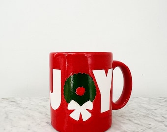 May include: A red ceramic mug with the word "JOY" in white lettering. A green wreath with a red center and white bow is in the center of the word "O". The mug has a red handle and is on a marble surface.