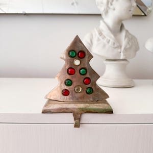 May include: A silver-toned Christmas tree stocking holder with red, green, and clear jewel-toned accents. The tree-shaped holder has a hook for hanging stockings. The design is festive and decorative, perfect for holiday decor.