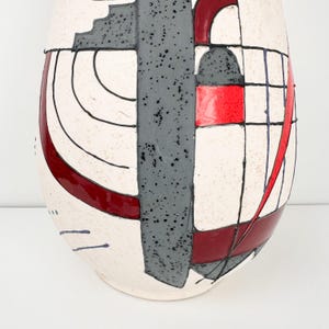 May include: A tall, abstract ceramic vase with a cream-colored base. The vase features a geometric design with red, gray, and black accents. The design includes squares, rectangles, and curved lines, creating a modern aesthetic.