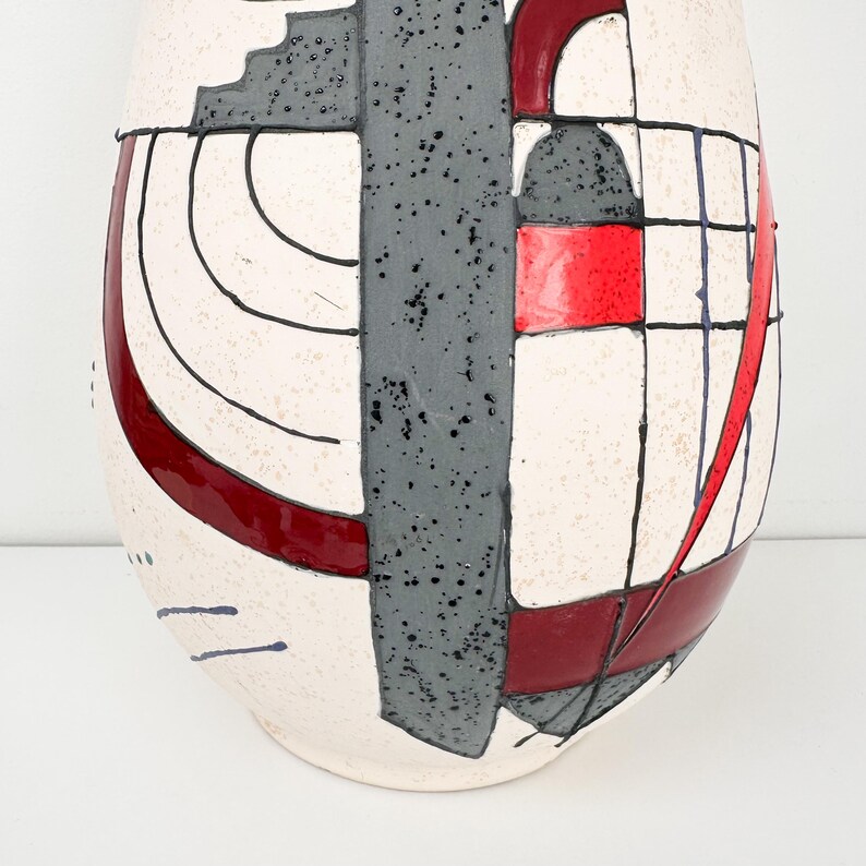 May include: A tall, abstract ceramic vase with a cream-colored base. The vase features a geometric design with red, gray, and black accents. The design includes squares, rectangles, and curved lines, creating a modern aesthetic.