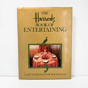 May include: A gold book with the title "The Harrods Book of Entertaining" by Lady Macdonald of Macdonald. The cover features a photo of a beautifully set table with a candle in the center.