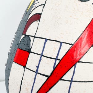 May include: A ceramic vase with a unique, abstract design. The vase features a white base with a grid pattern of blue lines and black outlines. Bold red and gray shapes add contrast. The design is reminiscent of modern art.