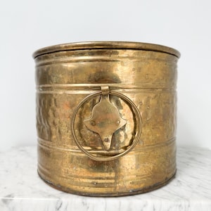 May include: A cylindrical brass bucket featuring a hammered texture on the lower section and a smooth upper band. The container includes a decorative brass plate with a circular ring handle attached to the side, showing a weathered patina.