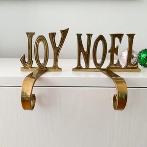 May include: Two polished brass stocking holders featuring the words JOY and NOEL in a stylized serif font. Each piece has a flat base with a curved hook extending downward, designed to hang from a ledge or mantelpiece with a metallic finish.