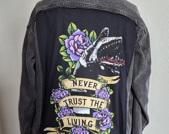 Könnte beinhalten: Graue Jeansjacke mit schwarzem Rückenteil, das eine Hai-Grafik mit offenem Maul, umgeben von lila Rosen und grünen Blättern, zeigt. Der Text "NEVER TRUST THE LIVING" ist auf einem Banner zu sehen.