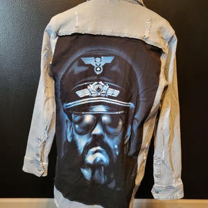 May include: Light blue denim jacket with a black t-shirt patch featuring a graphic of a man wearing a military cap and sunglasses. The jacket has distressed details and a frayed hem.
