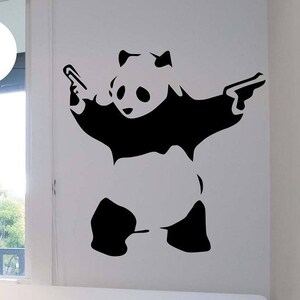 Puede incluir: Pegatina de pared en blanco y negro de un oso panda sosteniendo dos pistolas. La pegatina está en una pared blanca sobre una cama con una almohada a rayas azules y blancas. El texto "State Of The Wall" es visible en la imagen.