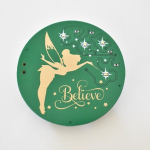 May include: A green circular ornament with a gold fairy silhouette and the word "Believe" in gold script. The fairy is holding a wand and there are small gold stars scattered around the ornament.
