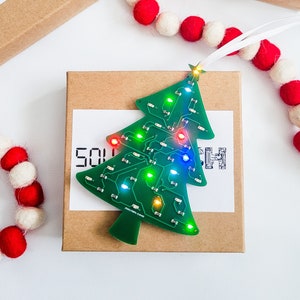 Op de afbeelding: Een groene kerstboom-versiering van een printplaat met kleurrijke LED-lampjes. De versiering ligt op een bruine doos met het woord "SOU" zichtbaar.