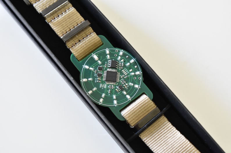 May include: A green circuit board with a black chip and silver components is attached to a beige nylon watch strap. The watch is in a black box.
