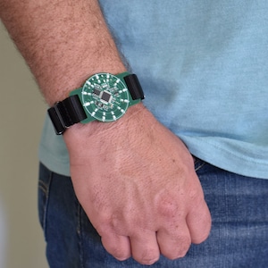 May include: A green circuit board with black wiring attached to a black wristband. The circuit board has a small integrated circuit chip in the center.