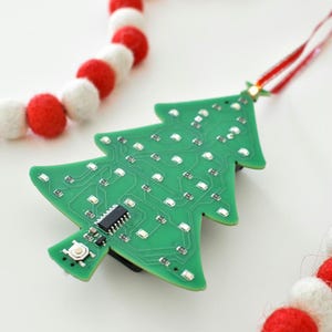 May include: A green circuit board Christmas tree ornament with LED lights. The tree-shaped ornament has a red and white ribbon for hanging. A red and white felt ball garland is in the background.