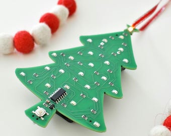 May include: A green circuit board Christmas tree ornament with LED lights. The tree-shaped ornament has a red and white ribbon for hanging. A red and white felt ball garland is in the background.