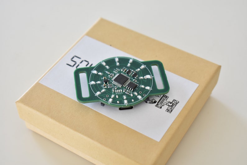 May include: A green circuit board with a circular design and a rectangular buckle. The board has a small chip in the center and several LEDs around the edge. The board is sitting on a brown box with a white label that says "SOUCH".