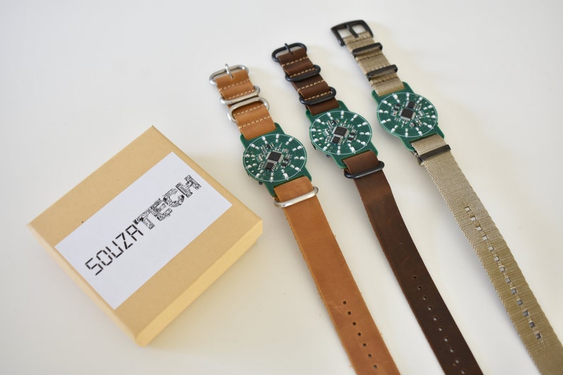 May include: Three wristwatches with green circuit board faces and different colored leather and fabric bands. The watches are arranged on a white surface with a small brown box labeled "SOUZA TECH" in the lower left corner.