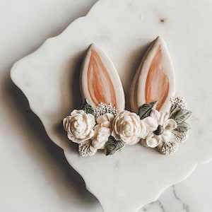 Puede incluir: Un par de orejas de conejo marrones con detalles florales blancos y verdes. Las orejas están hechas de pasta de azúcar y están decoradas con flores blancas y hojas verdes.
