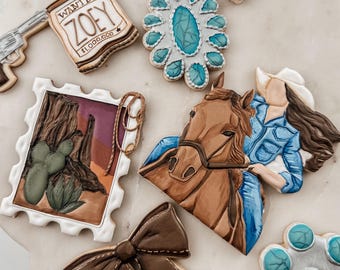 May include: Assortment of decorated sugar cookies with a Western theme. Designs include a "Wanted" poster, a horse and rider, a decorative bow, and a turquoise-colored floral design. The cookies are arranged on a marble surface.