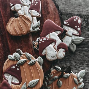 May include: An assortment of autumn-themed cookies, including pumpkins and mushrooms. The cookies feature brown, white, and burgundy colors with intricate details. The cookies are arranged on a wooden surface, creating a rustic aesthetic.