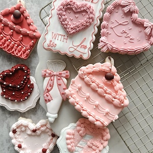 May include: A selection of decorated biscuits in heart and cake slice shapes. The biscuits are mainly pink and red, with white icing details and decorative elements. Some biscuits feature the words "Love" or "Baked with Love".