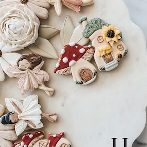 May include: A collection of intricately decorated sugar cookies featuring fairy and mushroom house themes. The cookies are decorated with delicate details, including flowers, leaves, and whimsical faces. The cookies are arranged on a white surface with the text "Jolano cookie cutters" in black.