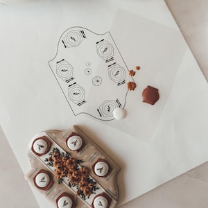 May include: A detailed overhead shot features a decorative cookie with a floral design and six circular elements, each with a sprig. A drawing of a table setting with plates and cutlery is also visible, along with a few small decorative items.