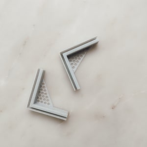 Puede incluir: Dos escuadras de esquina de plástico gris con un patrón de panal. Las escuadras están diseñadas para reforzar las esquinas y proporcionar soporte.