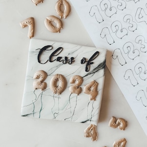 Può includere: Una torta bianca con la scritta "Class of" in lettere nere e numeri di palloncini dorati che compongono "2024". La torta è su una superficie con motivo a marmo. Ci sono altri numeri di palloncini dorati sparsi intorno alla torta.