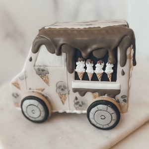 May include: A white ice cream truck with a brown chocolate glaze dripping down the sides. The truck has a window with four ice cream cones inside. The truck is sitting on a white marble surface.